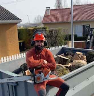 Un élagueur en tenue orange assis sur une remorque remplie de bois coupé, tronçonneuse en main